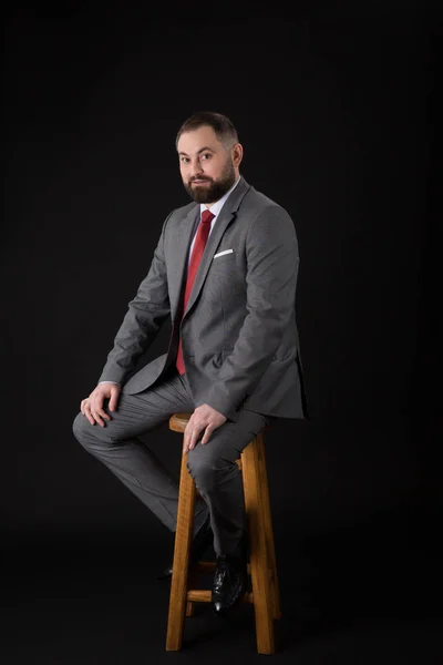 수염을 기고한 패션맨이 높은 의자에 앉아 카메라를 바라보며 양복을 입고 있는 모습. 검은 색 배경에 — 스톡 사진