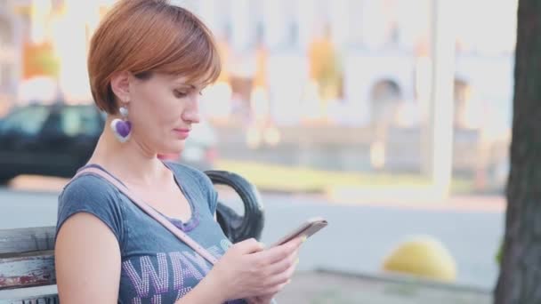 女人使用智能手机放松在长凳上在街上。慢动作，4k。年轻的千禧年妇女在植物园在电话显示屏上做手势. — 图库视频影像