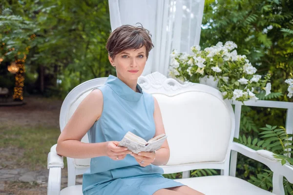 summer garden, good mood, retro style. adult Dreamy brunette of fifty sitting on a romantic white vintage bench with a book in her hands.