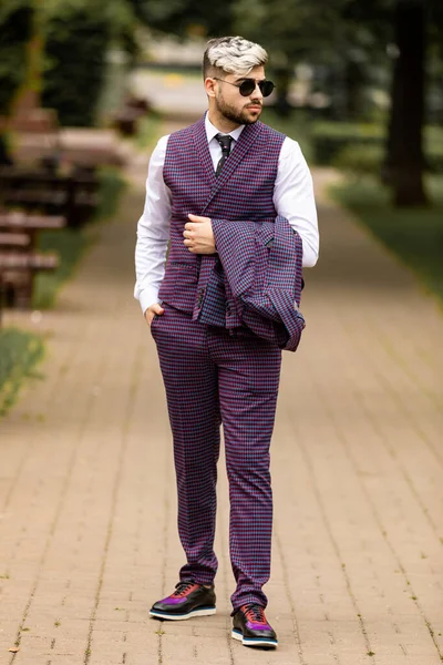 サングラスをかけて紫の高級3ピーススーツを着た若いビジネスマンが通りを歩いていると横に見える 上着を手に持った男が — ストック写真