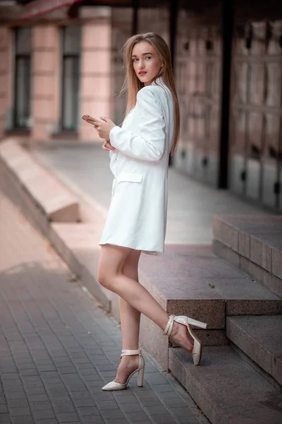 Retrato de hermosa joven mujer de negocios con teléfono en la mano en la calle de verano. Modelo femenino con el pelo largo en traje blanco de pie en la ciudad —  Fotos de Stock