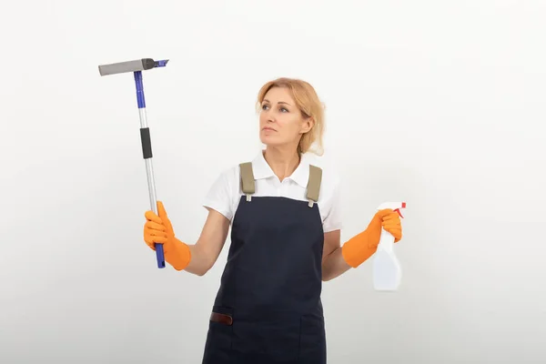 Portrait of cleaning lady or housewife holding a window cleaning brush and a spray. Wearing a uniform apron and latex gloves on light background. European female cleaner ready to work. cleaning agency provides its services