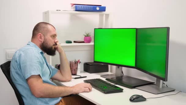 Un uomo che lavora a casa davanti a due monitor verdi. un uomo pensa e ha un'idea. — Video Stock