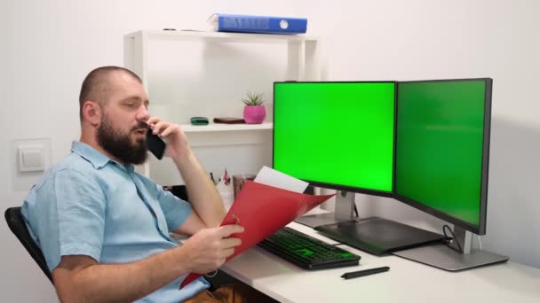 Człowiek zagląda do folderu i rozmawia przez telefon. krzyczy na rozmówcę. człowiek pracujący w domu przed dwoma zielonymi monitorami. — Wideo stockowe