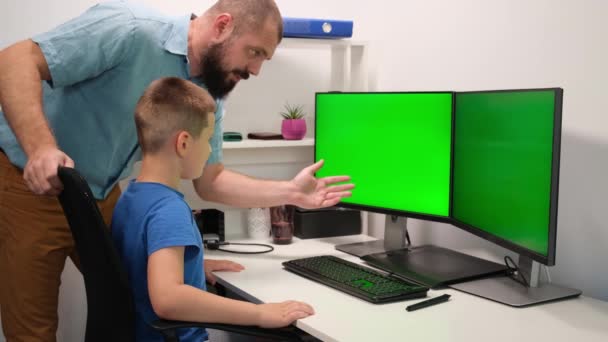 Padre e hijo hacen tareas remotamente frente a dos monitores verdes. el hombre explica el nuevo material al niño. — Vídeos de Stock