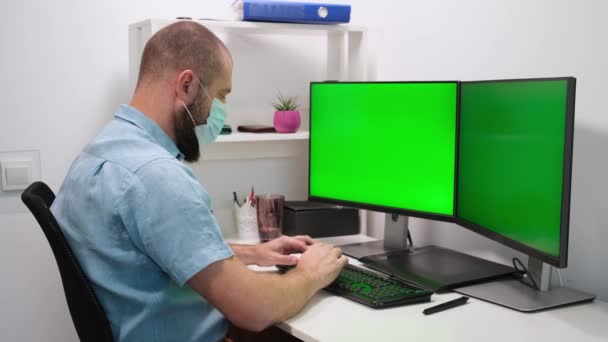 Hombre de negocios freelancer que trabaja en la computadora en casa. Dos pantallas verdes. Un hombre con una máscara médica trabaja delante de dos monitores verdes. Trabajo remoto en casa en cuarentena de autoaislamiento — Vídeos de Stock