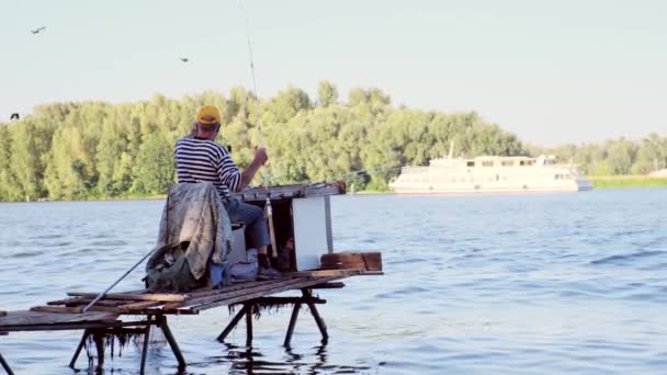 Een man met een hengel op de rivier is aan het vissen. Een visser met een draaiende hengel vist op een houten platform. Visser op de achtergrond van de rivier, motorschip en blauwe lucht. — Stockvideo