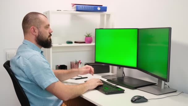 Manos de hombre barbudo escribiendo en el teclado en dos monitores con la pantalla verde — Vídeos de Stock