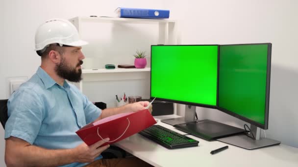 Ingeniero que usa casco de seguridad o Hardhat funciona en una computadora, dos pantallas de monitor muestran pantallas verdes. trabajo remoto en casa — Vídeo de stock
