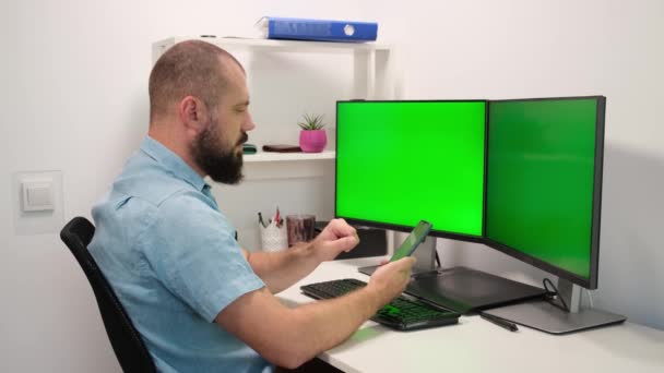 Hombre sentado en casa y mensajes de texto en el teléfono inteligente contra dos pantallas verdes o monitores. Dedos escribiendo en la pantalla táctil — Vídeos de Stock