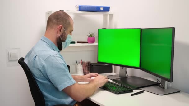 Άνθρωπος με ιατρική μάσκα πληκτρολογώντας στο πληκτρολόγιο μπροστά σε δύο πράσινες οθόνες. εξ αποστάσεως εργασία στο σπίτι σε καραντίνα — Αρχείο Βίντεο