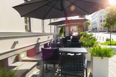 Şehir caddesinde boş bir kafeterya terası. Masaları, sandalyeleri ve büyük siyah şemsiyeleri olan. Yaz günü güneş ışığıyla yıkanmış.