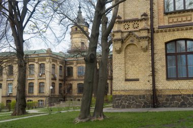 Kyiv. Kyiv Politeknik Enstitüsü. Geniş, sarı tuğla bina, detaylı mimari özelliklere sahip, sayısız pencere, ve bulutlu bir günde çevreleyen yeşil çimenler ve ağaçlar.
