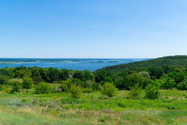 Uçsuz bucaksız bir nehrin büyüleyici panoramik manzarası yemyeşil ormanlar ve yuvarlanan tepeler boyunca akar, açık mavi gökyüzünün altında ufka uzanır.
