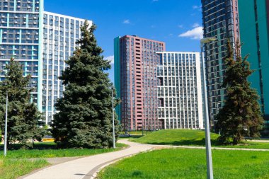 Parlak mavi bir gökyüzü altında, uzun, modern yerleşim binalarıyla çevrili, yemyeşil çimenli ve yemyeşil ağaçları olan canlı bir yeşil park.
