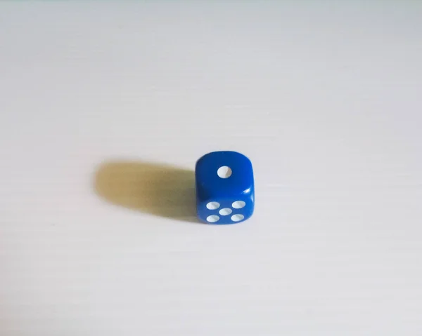 Sade bir ışık yüzeyindeki tek bir mavi zarın yakın plan fotoğrafı. Zarlar, solunda yumuşak bir gölgeyle üstteki bir numarayı gösteriyor. Minimalist kompozisyon basitliği vurgular ve oyun nesnesine odaklanır.