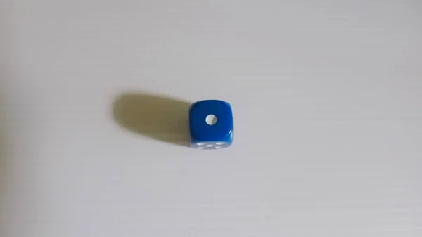 Sade bir ışık yüzeyindeki tek bir mavi zarın yakın plan fotoğrafı. Zarlar, solunda yumuşak bir gölgeyle üstteki bir numarayı gösteriyor. Minimalist kompozisyon basitliği vurgular ve oyun nesnesine odaklanır.