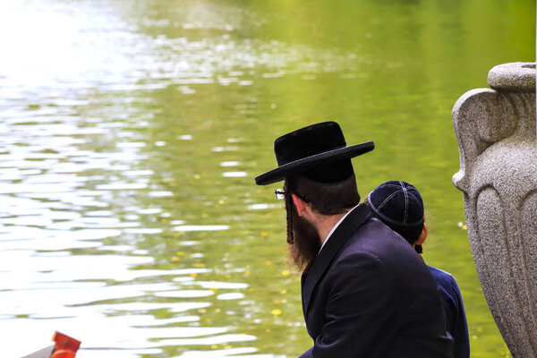 A family of Hasidic Jews, a Jewish man with a boy, in traditional black clothes, are sitting by the lake in a park, Uman, Ukraine