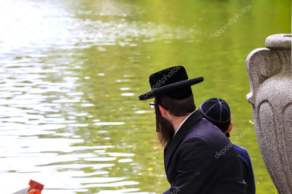 A family of Hasidic Jews, a Jewish man with a boy, in traditional black clothes, are sitting by the lake in a park, Uman, Ukraine