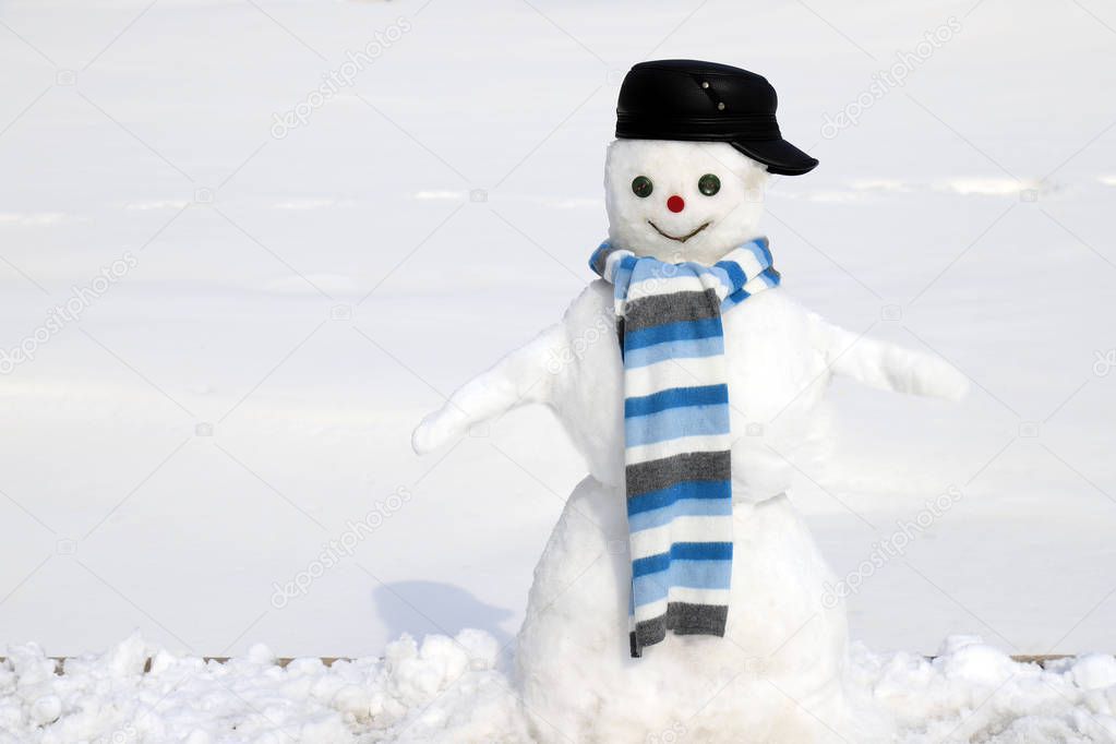 A snowman in a black cap and a blue scarf is standing on a snowy meadow in winter, Christmas and New Year holidays, entertainment and sports.