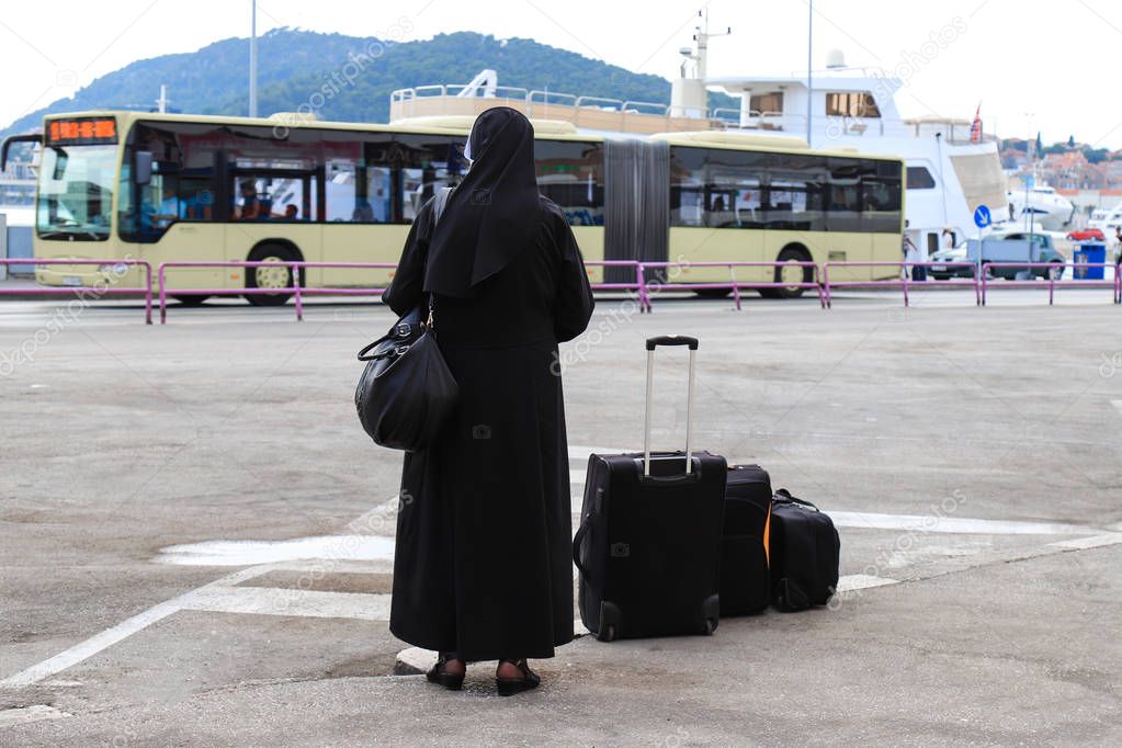 Nun traveling with suitcases and luggage. A woman in black monastic clothes is waiting for a bus on the waterfront of the old Croatian city Split. Travel in Croatia