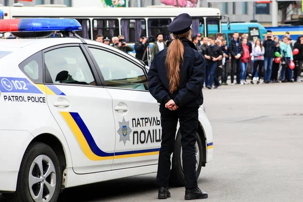 Дівчина – поліцейський, що стоїть біля патрульної поліцейської машини з написом українською мовою-патрульної поліції та Герб України — стокове фото