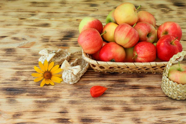 Red apples and autumn flowers lie in a wicker plate on a wooden table. Wicker bast shoes and apples in a basket. Healthy food and lifestyle, vegetarianism. — Stock Photo, Image
