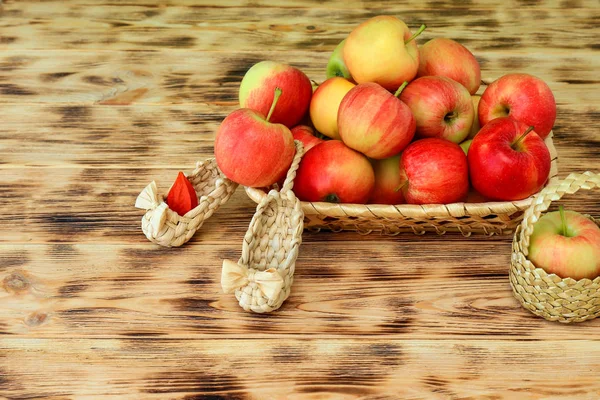 Rote Äpfel und Herbstblumen liegen in einem Weidenteller auf einem Holztisch. Weidenschuhe und Äpfel in einem Korb. gesunde Ernährung und Lebensweise, Vegetarismus. — Stockfoto