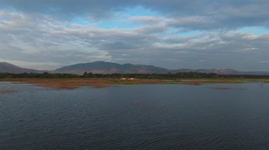 Orman tepeleri ve dağlarla desteklenen uzak sulak arazili sakin mavi su engin bulutlarla dolu gökyüzü - 4K yatay