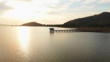 Sakin göl, yumuşak günbatımı ışığı altında parıldıyor. Küçük bir kulübeye ve uzak tepelere giden uzun bir iskele var. 4K yatay.