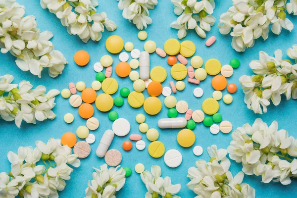 Різнокольорові таблетки лежать на синьому фоні, оточені білими суцвіттями акації, концепція алергії — стокове фото