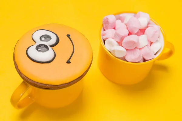 Två gula koppar på gul bakgrund, smiley-formade kakor ovanpå koppen, en annan kopp full av marshmallow — Stockfoto