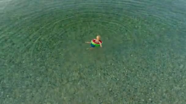 Маленький улыбающийся мальчик с высоким углом плавает в чистой воде, используя спасательный круг в летний вечер. — стоковое видео