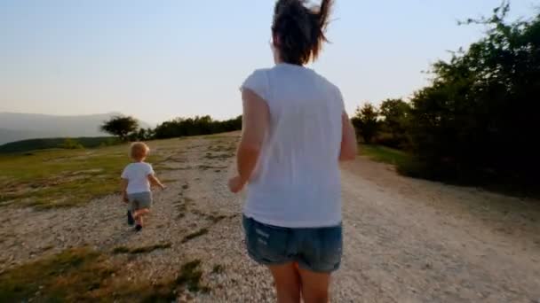 Feliz joven madre e hijo pequeño corriendo a lo largo del camino rural hacia el atardecer cámara lenta — Vídeos de Stock