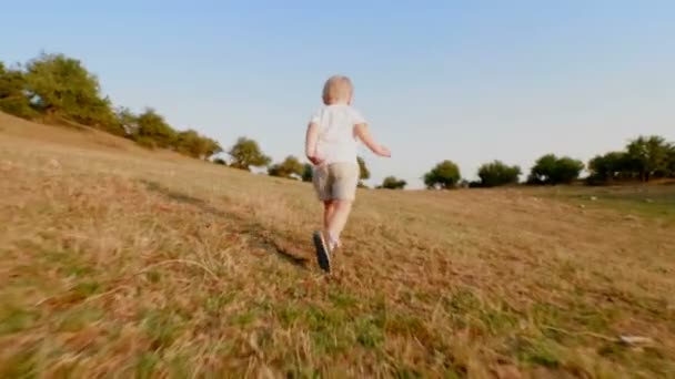 Niño feliz corriendo en el prado con hierba amarilla en terreno montañoso al atardecer bajo ángulo — Vídeos de Stock