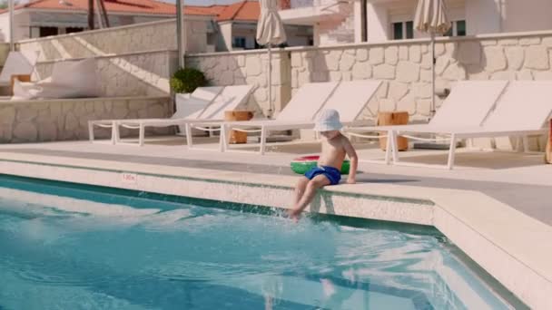 Lite aktiv turist pojke bär hatt leende och spelar ben sitter på kanten vid poolen — Stockvideo