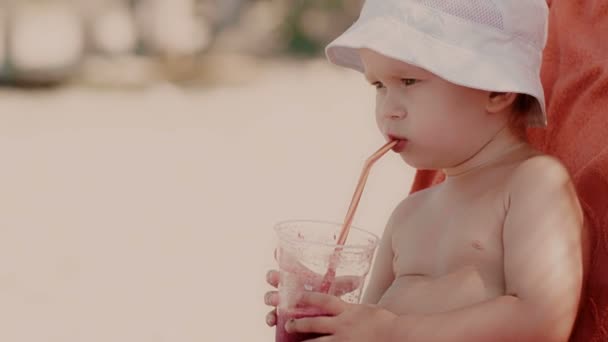 Retrato de un niño pequeño con sombrero frunciendo el ceño y bebiendo jugo fresco usando paja en la playa — Vídeo de stock