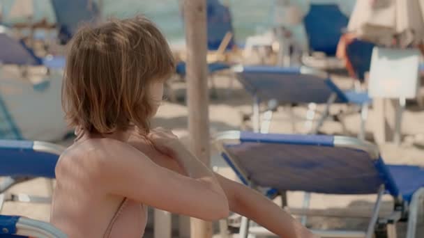 Yan görünümü turist çekici genç kadın vücudunda şezlong üzerinde oturan güneş koruyucu krem uygulamak — Stok video