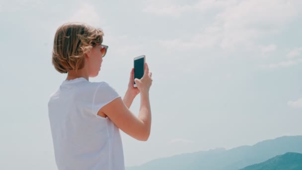 Hermosa chica joven con el pelo ondulado haciendo foto usando el teléfono inteligente cielo azul claro en el fondo — Vídeos de Stock