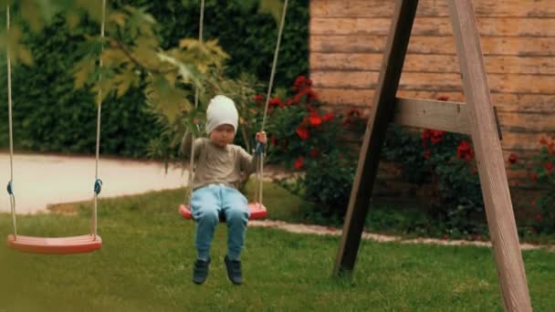 Niño casual feliz relajándose balanceándose en el columpio pasándolo bien en el patio de recreo cámara lenta — Vídeos de Stock