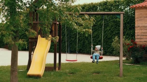 Aktiv liten söt baby boy swinging på Swing på lekplats omgiven av gröna träd — Stockvideo