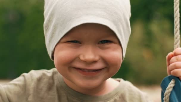 Крупным планом улыбающееся лицо маленького милого малыша во время раскачивания на качелях, позирующее глядя в камеру — стоковое видео