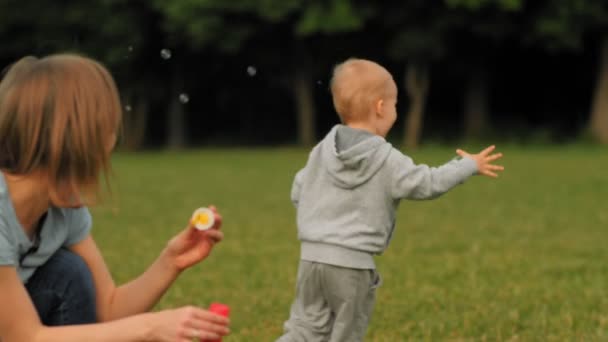 Улыбающаяся молодая мать надувает мыльный пузырь, играя с маленьким милым сыном на зеленой траве — стоковое видео