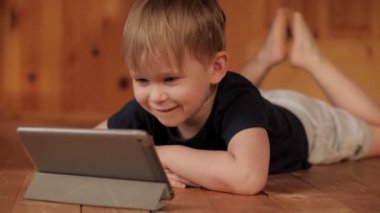 Neşeli çıplak ayaklı erkek çocuk ahşap zeminde uzanıyor rahatlıyor tablette eğlence videosu seyrediyor.