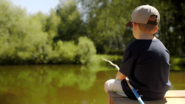 Ragazzo solitario seduto sul molo con canna da pesca vista posteriore pieno colpo carino pescatore bambino in attesa cattura — Video Stock