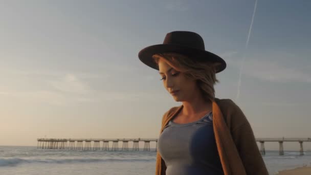 Красива дівчина в капелюсі грає пальто в вітряну погоду на пляжі на заході сонця крупним планом позує на відкритому повітрі — стокове відео