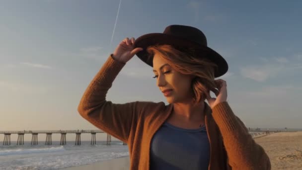 Atractiva joven viajera poniéndose un sombrero de moda relajándose en la playa al atardecer de cerca — Vídeos de Stock