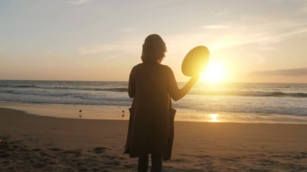 リラックスした女の子取りますオフ帽子遊び髪賞賛海に夕日バックビューパンニングショットスローモーション — ストック動画