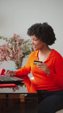 Çevrimiçi alışverişe katılan neşeli genç bir kadın, dizüstü bilgisayarına odaklanırken kredi kartını güvenle kullanıyor, evden mal almanın kolaylığını ve eğlencesini gösteriyor.