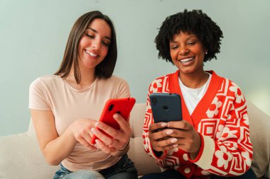 İki gülümseyen dişi koltukta otururken cep telefonlarını neşeyle kullanıyorlar. Heyecan verici bir dijital deneyimle meşguller. Uygulamaları paylaşıyorlar ve akıllı telefonlarıyla iletişim kuruyorlar.